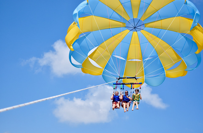 Parasailing in the Outer Banks is the perfect road trip adventure for all ages- blue skies with a blue and yellow parasailing with three happy people in the air!