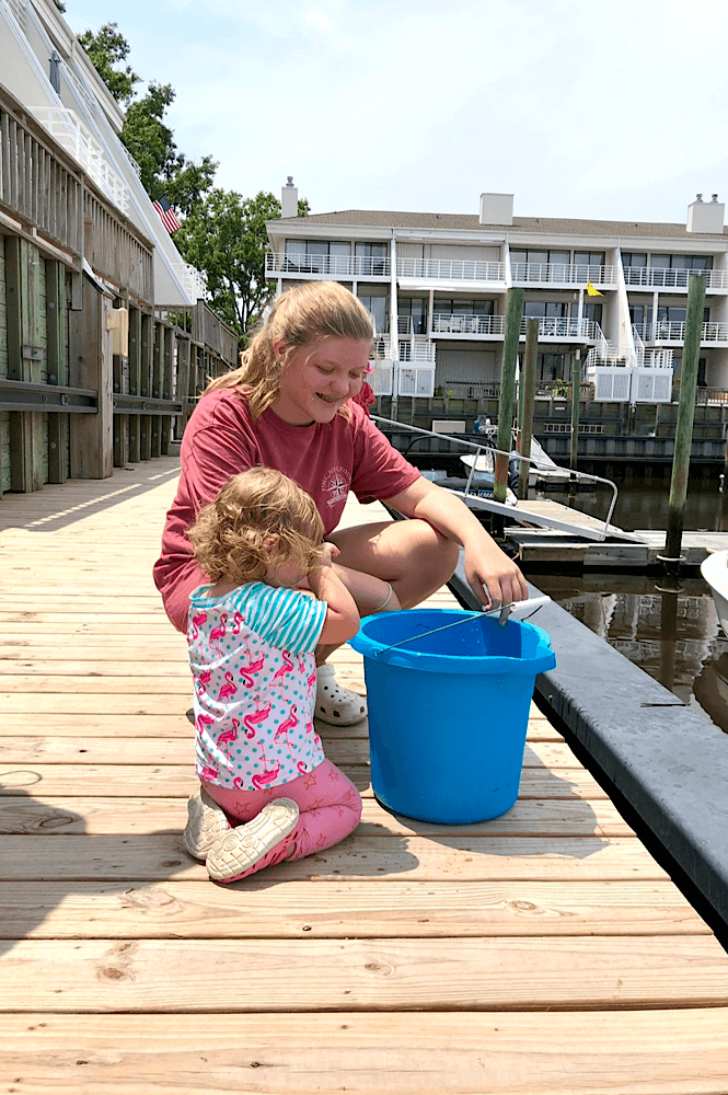 A toddler and a teenager looking at some fish in a bucket at a marina.