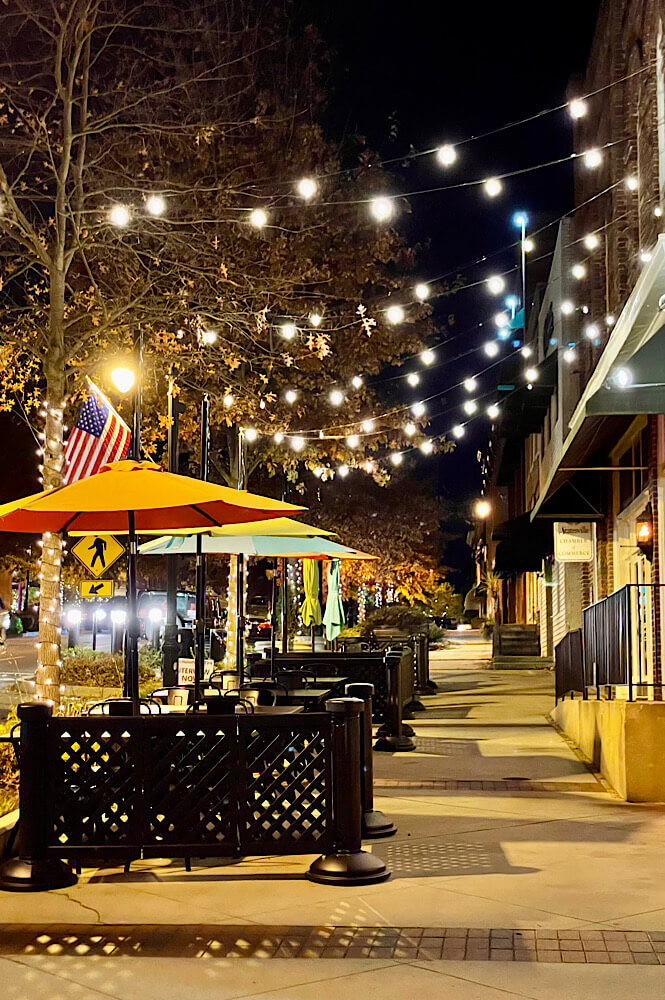 Tables with umbrellas in downtown Statesville NC at night lit up by string lights.