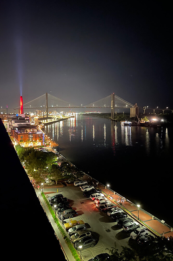 Nighttime city view of Savannah Georgia; the bridge in the background and the city is lit up with reflections on the river