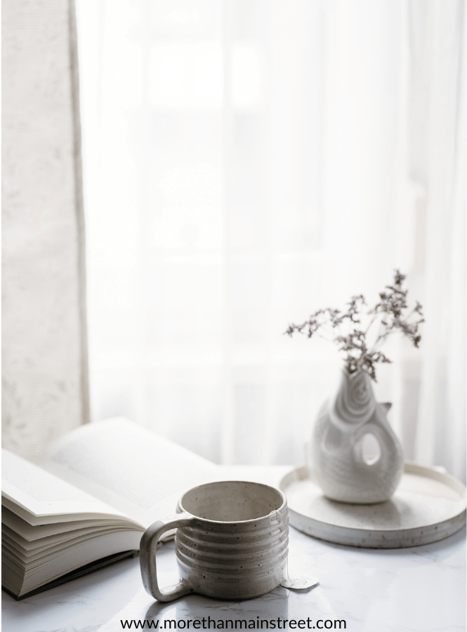 Slow living- image is an example of simple mornings. Image of a white desktop near a window with a book, coffee cup and small flower vase on it