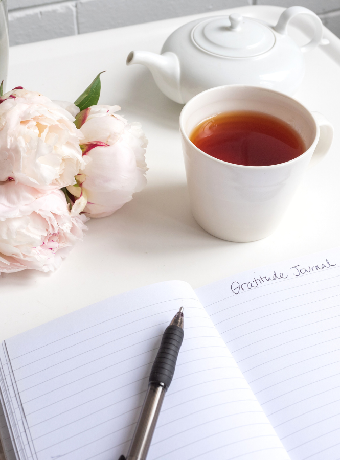 A cup of tea with a gratitude journal, pen, and flower on a white desk showing a slow intentional lifestyle