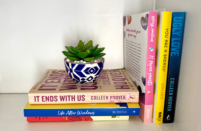 A stack of three books with a little succulent on top and three more books upright on a book shelf