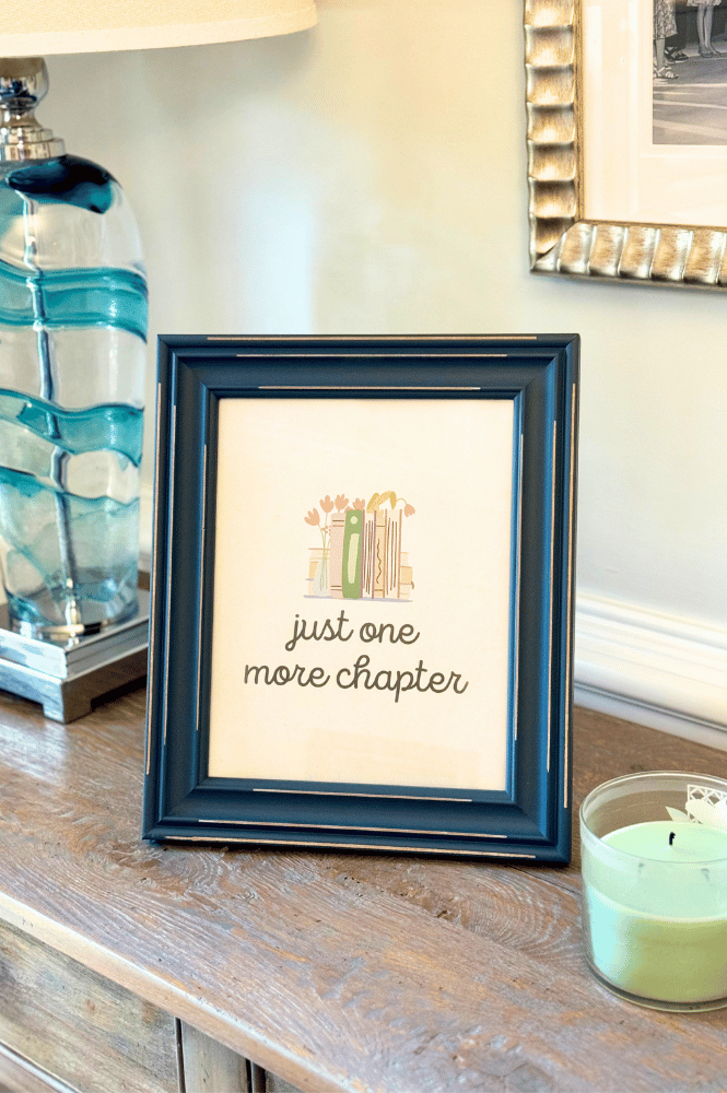 Free printable wall art- a printed framed paper that sawys "just one more chapter" on a side table with a lamp and candle
